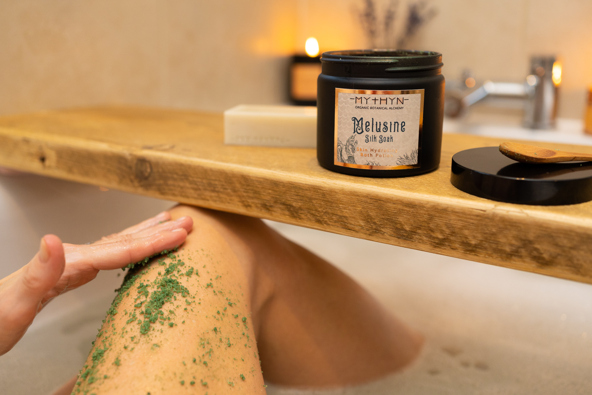 lifestyle product photography of bath soak scrub being massaged into skin in bath tub with a wooden bath board and candle in the background